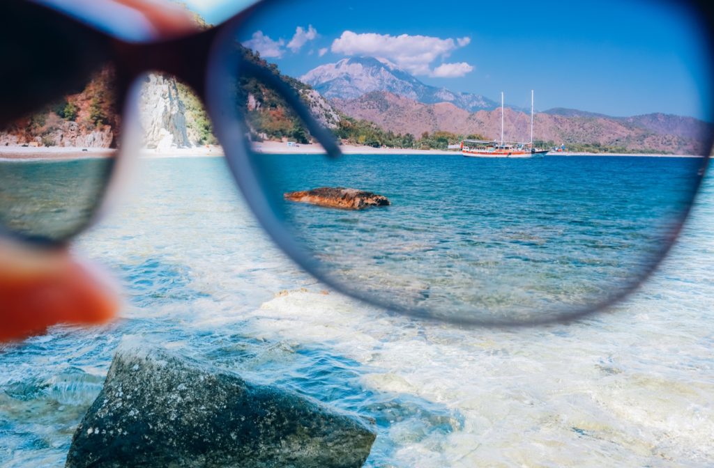A hand holding a pair of polarized glasses showing the effect with blue seawater, a boat, and mountains far in the background.